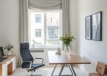 A modern home office featuring a large wooden desk, ergonomic chair, and a small wooden stool, with natural light coming through a window.