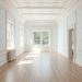 A spacious room with white wallpaper and a detailed ceiling, featuring large windows and light wood flooring.