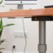 A modern adjustable desk with a wooden top and a sleek metal base, featuring a computer monitor and a power outlet in the background.
