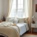 A cozy farmhouse bedroom featuring a bed with a chunky knit blanket, soft pillows, light curtains, and a woven rug.