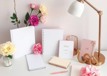 A feminine home office desk arrangement with notebooks, flowers, and a lamp.