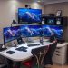 A modern tech-friendly workstation with three monitors, ergonomic chairs, and stylish LED lighting.