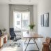 A modern home office featuring a large wooden desk, ergonomic chair, and a small wooden stool, with natural light coming through a window.